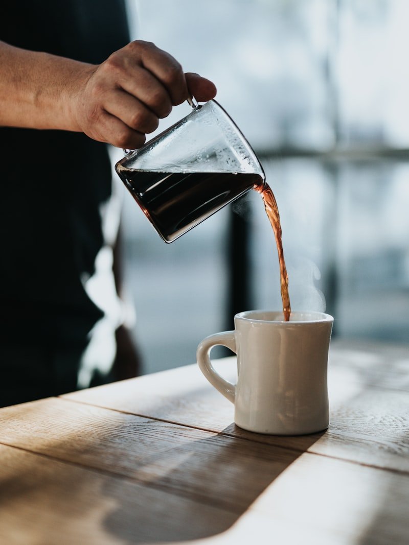 Mano sirviendo café en una taza sobre mesa de madera con luz de mañana